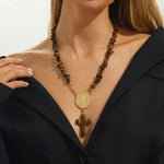 A close-up of a model wearing a gold-tone necklace with a round medallion and cross pendant, suspended from a strand of polished tiger's eye beads, styled with a black blazer.