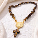 A beaded necklace featuring brown and black tiger's eye stones with a gold-tone Saint Benedict medal and cross pendant, displayed on a textured white fabric background.