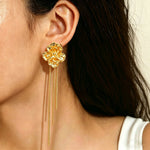 A close-up of a model's ear wearing a gold flower-shaped dangle earring with three long thin chains hanging below.