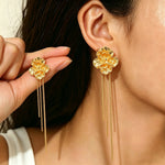 A close-up lifestyle shot of a model wearing a pair of gold flower-shaped dangle earrings, with one earring shown on her ear and the other held between her fingers.