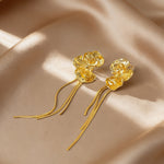 A pair of gold dangle earrings with textured flower-like tops and long thin dangling wires, displayed on draped satin fabric.