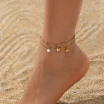 A close-up of a person's ankle and foot wearing two delicate gold anklets on sandy ground. One anklet has a small white pearl bead, and the other has a gold starfish and shell charm.