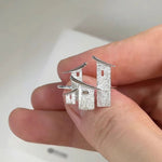 A close-up of a hand holding a pair of silver earrings shaped like textured Japanese torii gates.