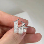 A close-up of a hand holding a pair of silver earrings shaped like stylized pagoda roofs or small architectural structures, shown against a soft light gray background.