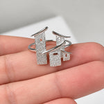 A silver ring with a textured, house-shaped design, shown resting on a person's fingers against a light background.