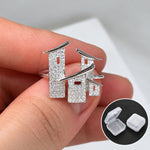 A close-up of a hand holding a pair of silver-toned house-shaped stud earrings with a textured finish. A small inset in the bottom right shows two clear plastic storage cases.