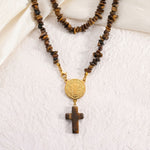 A beaded necklace featuring brown tiger's eye stones and a gold-tone Saint Benedict medallion with a matching wooden cross pendant, displayed on a textured white fabric background.