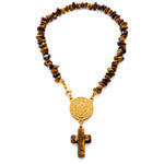 A beaded necklace with brown and black tiger's eye stones, a gold-tone Saint Benedict medallion, and a matching cross pendant, displayed on draped white fabric.